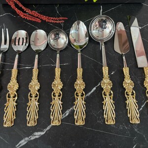 Op de afbeelding: Set van acht gouden serveerbestek, waaronder een vork, twee lepels, een soeplepel, een serveerlepel, een taartschep en twee botermesjes. Het bestek heeft sierlijke, decoratieve handvatten.