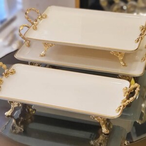 May include: Three cream-coloured rectangular serving trays with gold trim and ornate gold handles. The trays are supported by decorative gold feet. They are stacked on a reflective surface, suitable for serving food or displaying items.