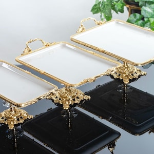 Puede incluir: Tres bandejas de servir de cerámica blanca con asas doradas y patas ornamentadas. Las bandejas están dispuestas en orden descendente de tamaño.