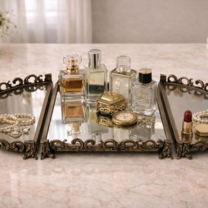Könnte beinhalten: Ein verzierter, verspiegelter Schminktisch-Tablett mit Parfümflaschen, Schmuck und einer kleinen goldenen Schatulle. Das Tablett hat einen dekorativen, bronzefarbenen Rahmen und steht auf einer Marmoroberfläche. Ein Strauß weißer Rosen ist im Hintergrund.
