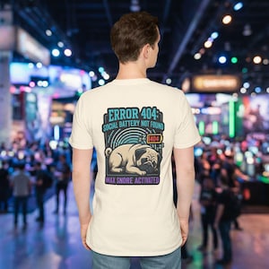 Könnte beinhalten: Ein cremefarbenes T-Shirt mit einem Aufdruck auf dem Rücken. Der Aufdruck zeigt einen schlafenden Mops mit dem Text "ERROR 404: SOCIAL BATTERY NOT FOUND" und "MAX SNORE ACTIVATED". Das Design ist in Blau-, Schwarz- und Beigetönen gehalten.