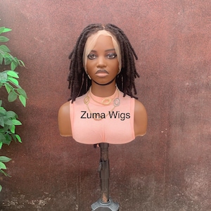 May include: A mannequin head displaying a dark brown dreadlock wig with a lace front. The mannequin is wearing a peach-colored top with the text "Zuma Wigs". The wig is styled on a black tripod stand.