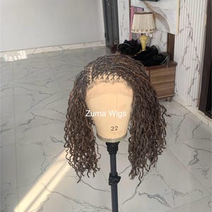 Puede incluir: Una peluca marrón y negra con textura rizada, exhibida en una cabeza de maniquí. La peluca tiene una línea de cabello de aspecto natural y está etiquetada como "Zuma Wigs" con el número "22". La peluca está peinada con rastas individuales.