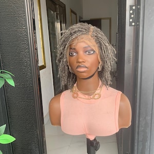 May include: A mannequin head displaying a gray dreadlock wig with a lace front. The mannequin is wearing a peach-colored tank top and a gold-toned necklace. The background includes a doorway and a glimpse of a room.