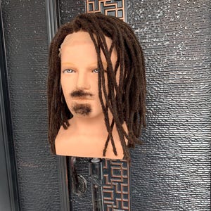 Op de afbeelding: Een bruin pruik met lange dreadlocks op een mannequinhoofd. De pruik is gestyled met een baard en snor.