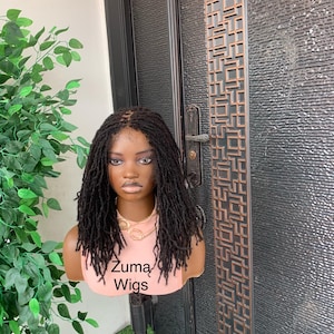 May include: A mannequin head displaying a black dreadlock wig. The mannequin is wearing a pink top and a gold necklace. The text "Zuma Wigs" is printed on the top. The wig is styled with individual dreadlocks. The background includes greenery and a dark door.