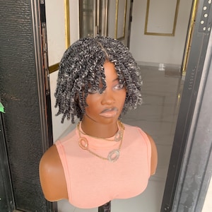 Peut inclure: Tête de mannequin avec une perruque courte torsadée grise et noire. Le mannequin porte un débardeur orange clair et un collier doré avec trois pendentifs décoratifs. L'arrière-plan comprend une porte et un mur avec des cadres dorés.