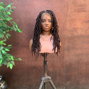 May include: A mannequin head with long, dark brown dreadlocks and a light brown hairline. The mannequin is wearing a pink top and a gold necklace. The head is mounted on a black tripod stand, set against a brown wall and green grass.