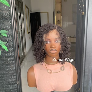 May include: A dark brown wig with a short, curly style is displayed on a mannequin head. The wig has a natural-looking hairline and is styled with a side part. The mannequin is wearing a peach-colored top and a gold necklace. The text "Zuma Wigs" is visible.