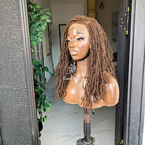May include: A brown wig featuring long, textured locs styled with a lace front base. The wig is displayed on a mannequin head mounted on a tripod stand. The hair has a natural, multi-tonal brown color with a matte finish and a defined center part.