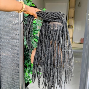 May include: A wig featuring long, black, textured dreadlocks styled on a mannequin head. The hair has a matte finish with a uniform, rope-like appearance throughout the length. The scalp area shows a defined parting with visible hair roots.