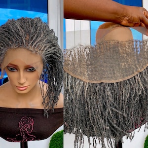Puede incluir: Una peluca de encaje frontal de cabello humano gris y negro con rastas. La peluca está en una cabeza de maniquí y el encaje es visible. La peluca está en un soporte con una camisa marrón debajo.