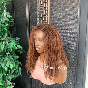 May include: A brown wig with a lace front, styled with small dreadlocks. The wig is displayed on a mannequin head, with the text "Zuma Wigs" visible on the image. The mannequin is wearing a pink top.