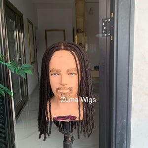 May include: A mannequin head displaying a dark brown dreadlock wig. The mannequin has a tan complexion and a beard. The text "Zuma Wigs" is visible. The wig features individual dreadlocks.