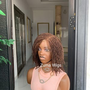 May include: A brown dreadlock wig on a mannequin head. The wig is styled with short, individual dreadlocks. The mannequin is wearing a pink top and a gold necklace with circular pendants. The text "Zuma Wigs" is visible.