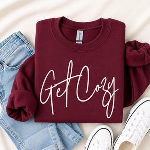 A folded maroon sweatshirt with 'Get Cozy' printed in white script across the front, styled in a flat lay with light blue jeans, white sneakers, and a beige fabric backdrop.