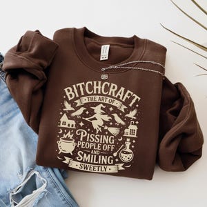 May include: A brown sweatshirt with the text "BITCHCRAFT the art of pissing people off and smiling sweetly" in a white graphic design. The design includes a witch on a broom, birds, and other witchy symbols. A silver necklace is draped over the sweatshirt.