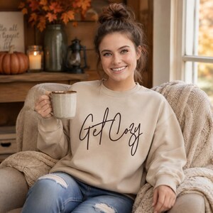 Woman wearing a sand Get Cozy sweatshirt while holding a mug in a warm fall living room.