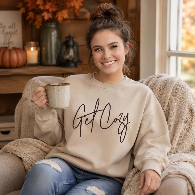 Woman wearing a sand Get Cozy sweatshirt while holding a mug in a warm fall living room.
