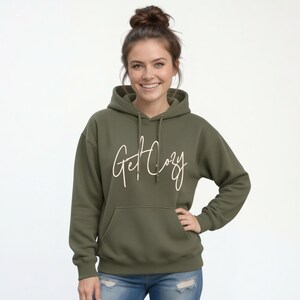 Woman wearing a military green Get Cozy hoodie with white script and jeans for a casual fall outfit.