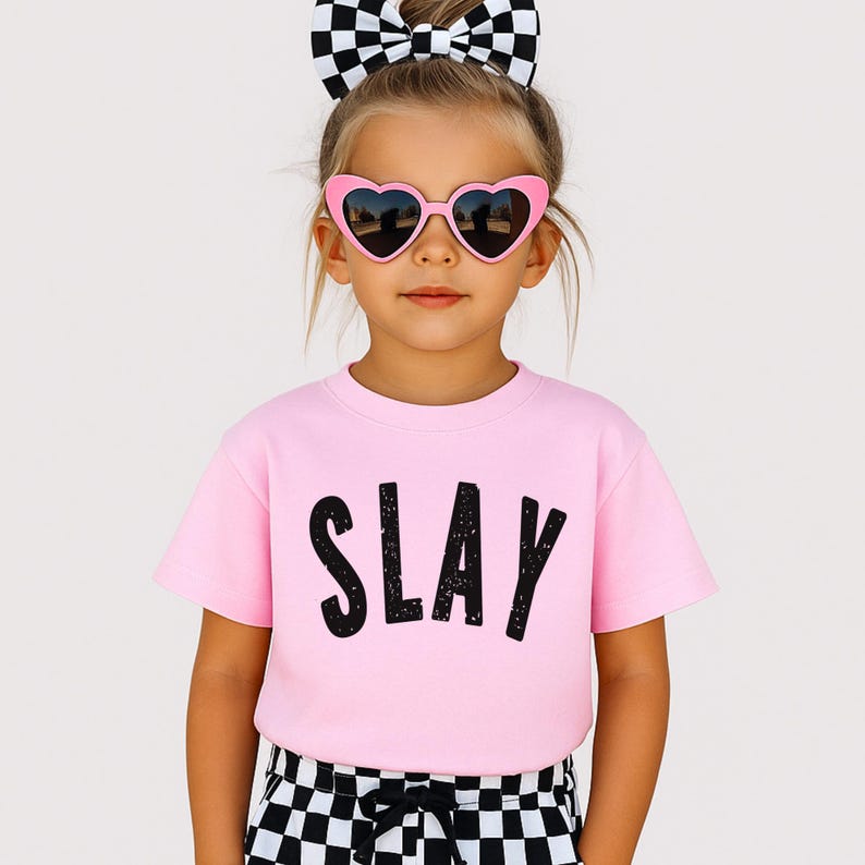 Adorable light pink SLAY graphic tee with bold black letters, modeled by stylish young girl in heart sunglasses. Youth sizing, perfect for tweens, back to school, matching with friends, or birthday gifts for confident girls who slay.