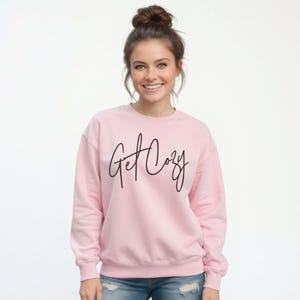 Woman wearing a light pink Get Cozy sweatshirt with black script and distressed jeans for a cozy fall outfit.