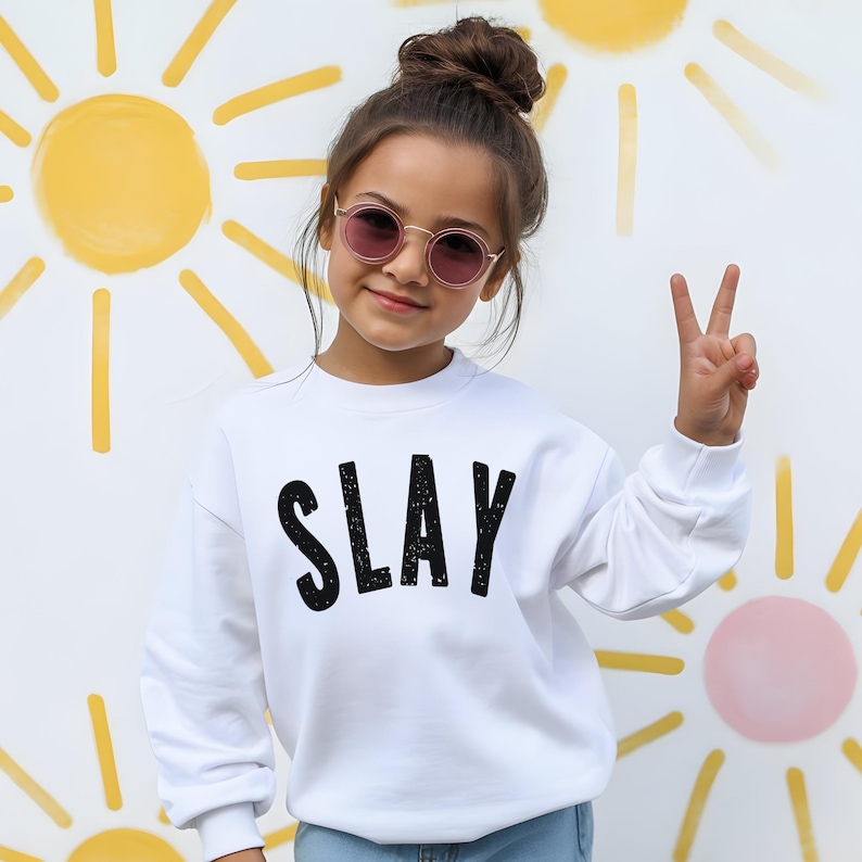 White youth sweatshirt with black SLAY graphic, girl wearing sunglasses giving peace sign, cute trendy kids outfit mockup, aesthetic girls clothing photo.
