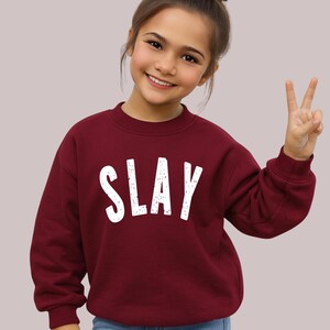 Maroon youth sweatshirt with white SLAY design, girl posing with peace sign, trendy kids aesthetic clothing mockup, cozy fleece crewneck for girls and teens.