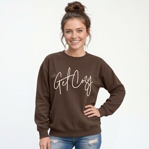 Woman wearing a brown Get Cozy sweatshirt with white script and jeans for a cozy fall outfit.