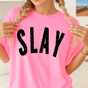 Bright pink SLAY T-shirt with bold black lettering, close-up view showing graphic design clearly. Trendy women’s or teen graphic tee mockup with cute braided hairstyle.