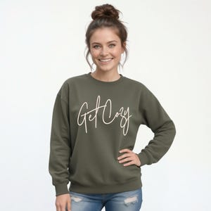 Woman wearing a military green Get Cozy sweatshirt with white script and distressed jeans for a cozy fall outfit.