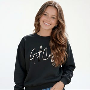 Girl wearing a black Get Cozy youth sweatshirt with white script and jeans.