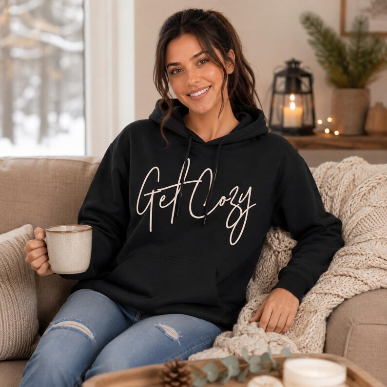Woman wearing a black Get Cozy hoodie while holding a mug in a cozy fall living room.