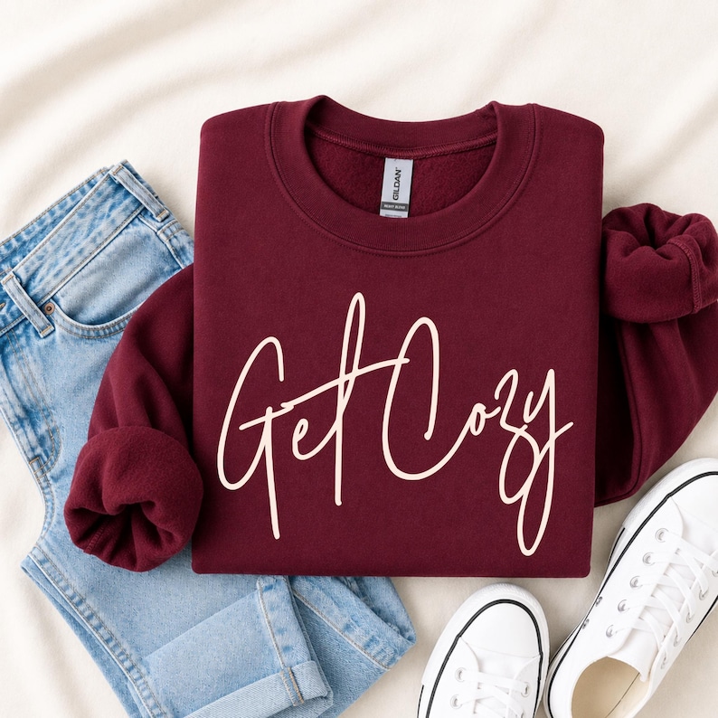 A folded maroon sweatshirt with 'Get Cozy' printed in white script across the front, styled in a flat lay with light blue jeans, white sneakers, and a beige fabric backdrop.