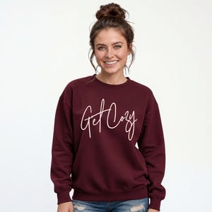 Woman wearing a maroon Get Cozy sweatshirt with white script and distressed jeans for a cozy fall outfit.