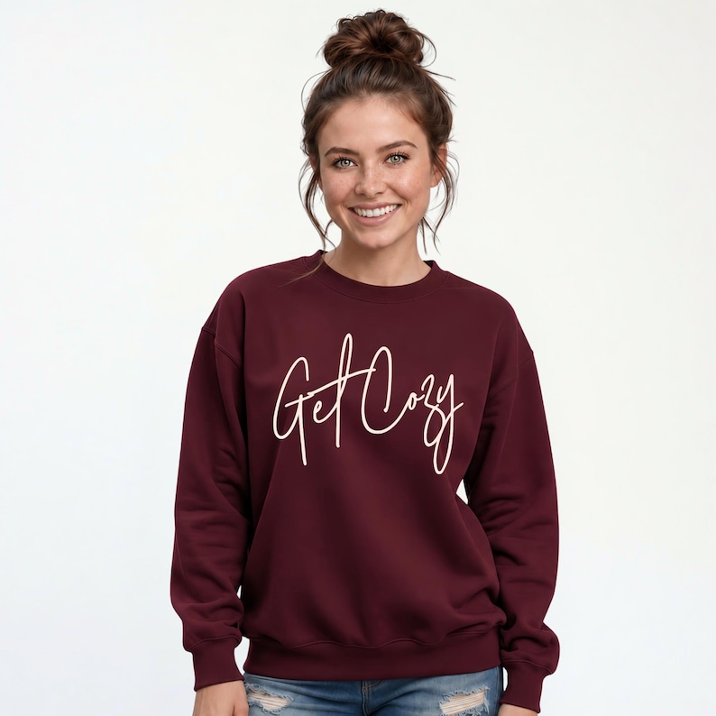 Woman wearing a maroon Get Cozy sweatshirt with white script and distressed jeans for a cozy fall outfit.