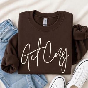 Brown Get Cozy sweatshirt flat lay with white script, light wash jeans and white sneakers.