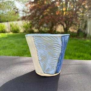 May include: A ceramic cup with a blue, white, and light blue glaze. The cup has a textured design and a rounded bottom.