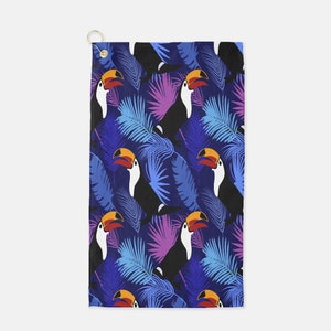 Puede incluir: Una toalla de golf con estampado azul y morado con tucanes y hojas de palma. Los tucanes tienen picos naranjas y cuerpos negros.