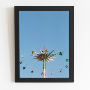 May include: A framed print featuring a carnival swing ride against a clear blue sky. The ride's central tower is green and white, with a starburst-shaped top in various colors. People are visible in the swings, adding a sense of motion.