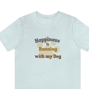 May include: A light blue t-shirt with the text 'Happiness is Running with my Dog' in dark blue and brown text. A paw print graphic is behind the text in a light brown color.