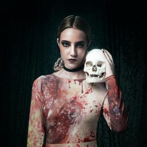 May include: A woman wearing a long-sleeved dress with a blood splatter pattern. She is holding a white skull in her right hand. The dress has a fitted bodice and a flowing skirt. The background is dark.