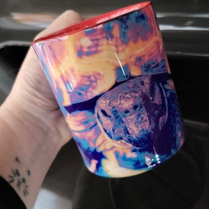 Könnte beinhalten: Eine bunte Keramiktasse mit einem Nahaufnahmebild eines Schildkrötenkopfes. Der Schildkrötenkopf ist scharf und der Hintergrund ist verschwommen. Die Tasse hat einen roten Rand und einen roten Griff.