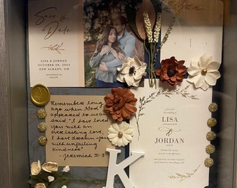 May include: A framed shadow box with a wedding invitation for Lisa and Jordan. The invitation is decorated with flowers, a handwritten quote, and a white letter K. The quote reads "Remember long ago when God appeared to us and said, "I have loved you with an everlasting love, I have dreamed your with unfailing kindness." - Jeremiah 31:3