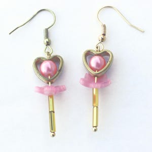 May include: A pair of dangle earrings with a heart-shaped frame and a pink pearl center. The earrings have a gold-colored hook and a gold-colored rod with pink accents and a small pink bead at the bottom. The earrings are on a white background.
