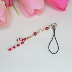 Puede incluir: Un charm de bastón de caramelo rojo y blanco unido a una cadena dorada con cuentas rosadas y blancas. La cadena está unida a una correa de teléfono negra.