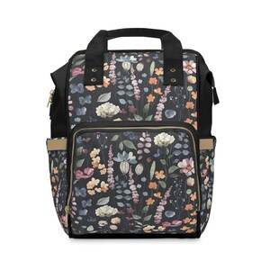 Op de afbeelding: Zwarte rugzak met een bloemenprint met aquarelbloemen in roze, oranje en blauwe tinten op een donkere achtergrond. De rugzak heeft een bovenste handvat en verstelbare schouderbanden.