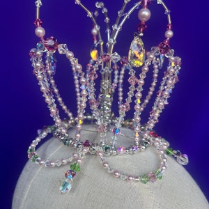 May include: A delicate crown made of silver wire and beads. The crown is adorned with pink and clear beads, and a large clear crystal in the center. The crown is sitting on a white mannequin head.