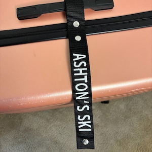 Puede incluir: Etiqueta de equipaje negra con el texto "ASHTON'S SKI" en blanco. La etiqueta está sujeta a una maleta rosa. La maleta tiene una cremallera y un asa negras.