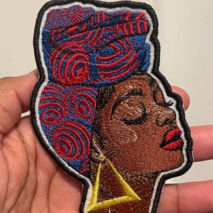 May include: Embroidered patch featuring a woman with her hair wrapped in a blue and red patterned head wrap. She is wearing a gold earring and has her eyes closed. The patch is outlined in black.